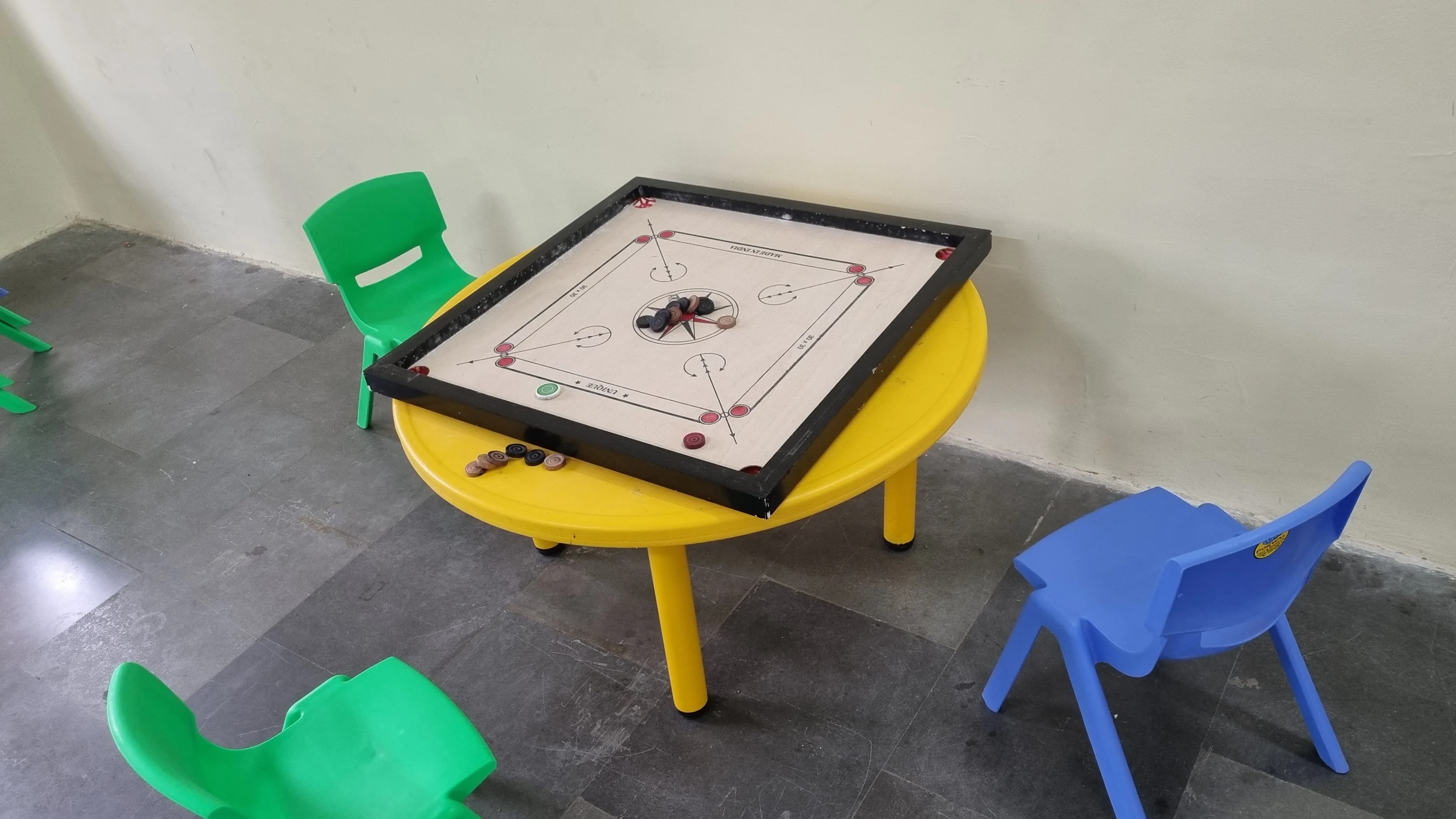 Carrom board on a yellow activity table surrounded by colourful chairs