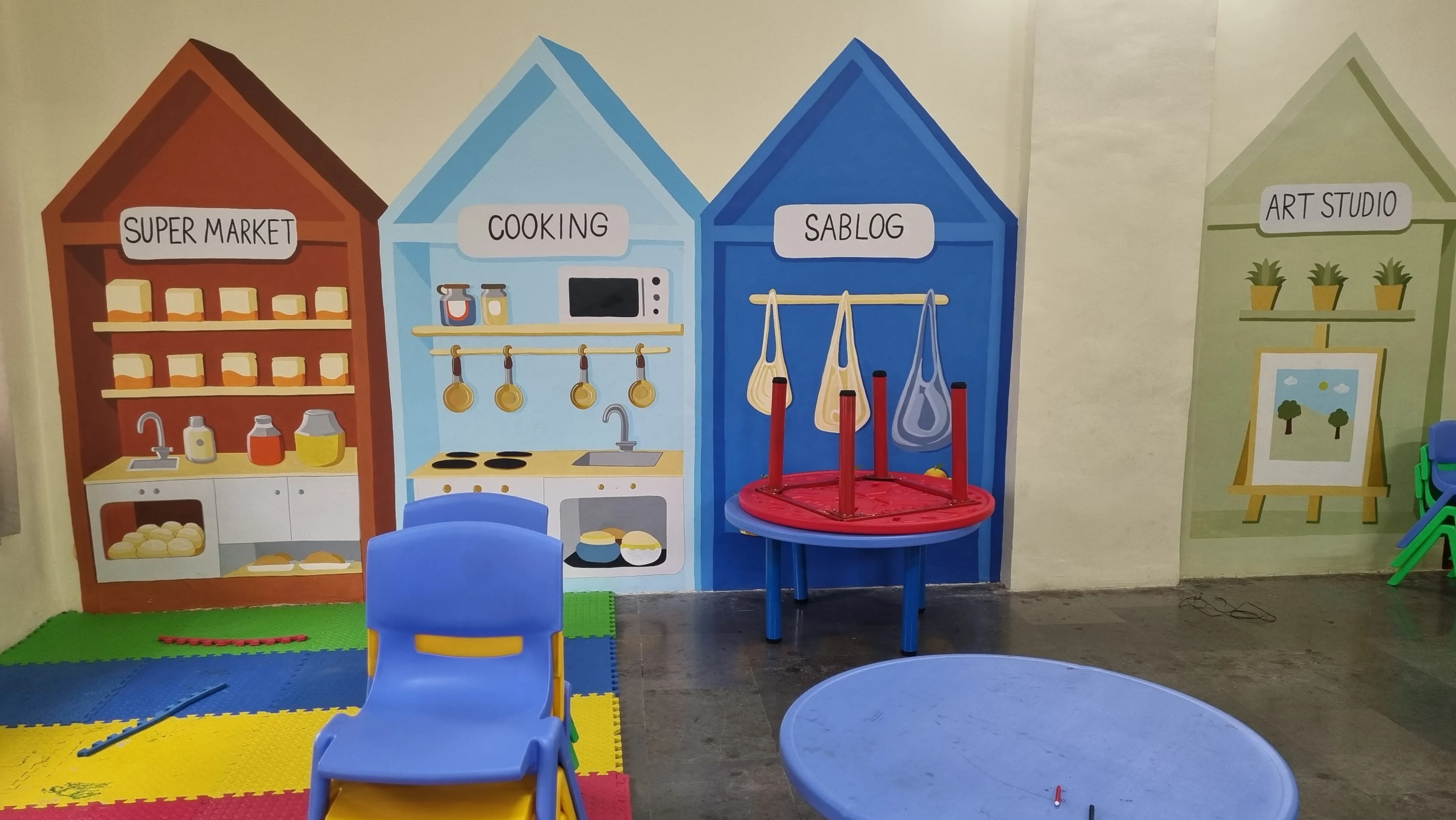 Colourful play-area mural showing a supermarket, kitchen, bag area and art studio